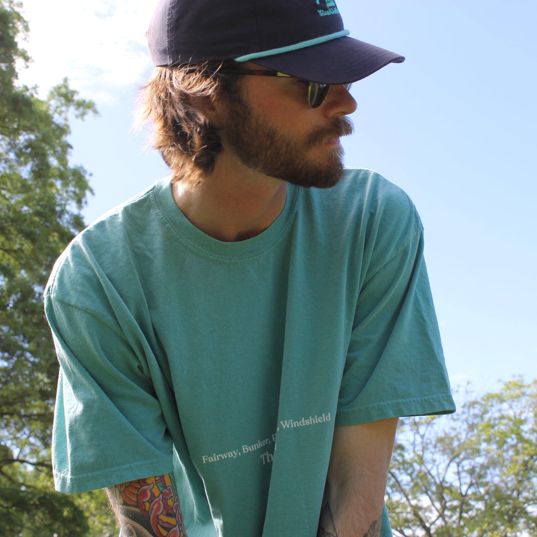 A golfer addresses his ball and looks at his target while wearing an aqua colored heavy cotton t-shirt with the expression "Fairway, bunker, pavement, windshield" on the front in small, centered text.