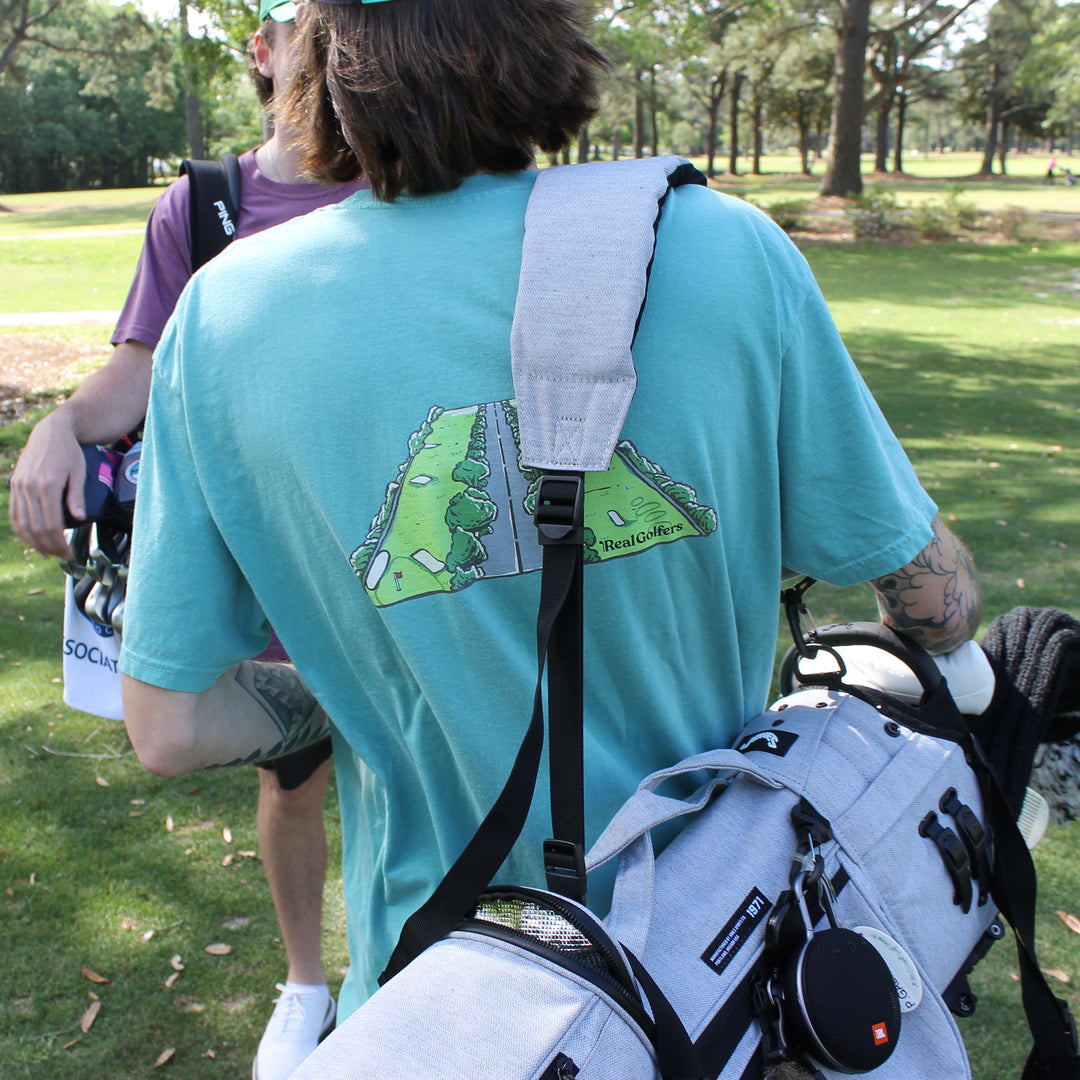 A golfer stands with his golf bag on his shoulder. He is wearing an aqua blue heavy cotton t-shirt with a design of a golf hole on the back.